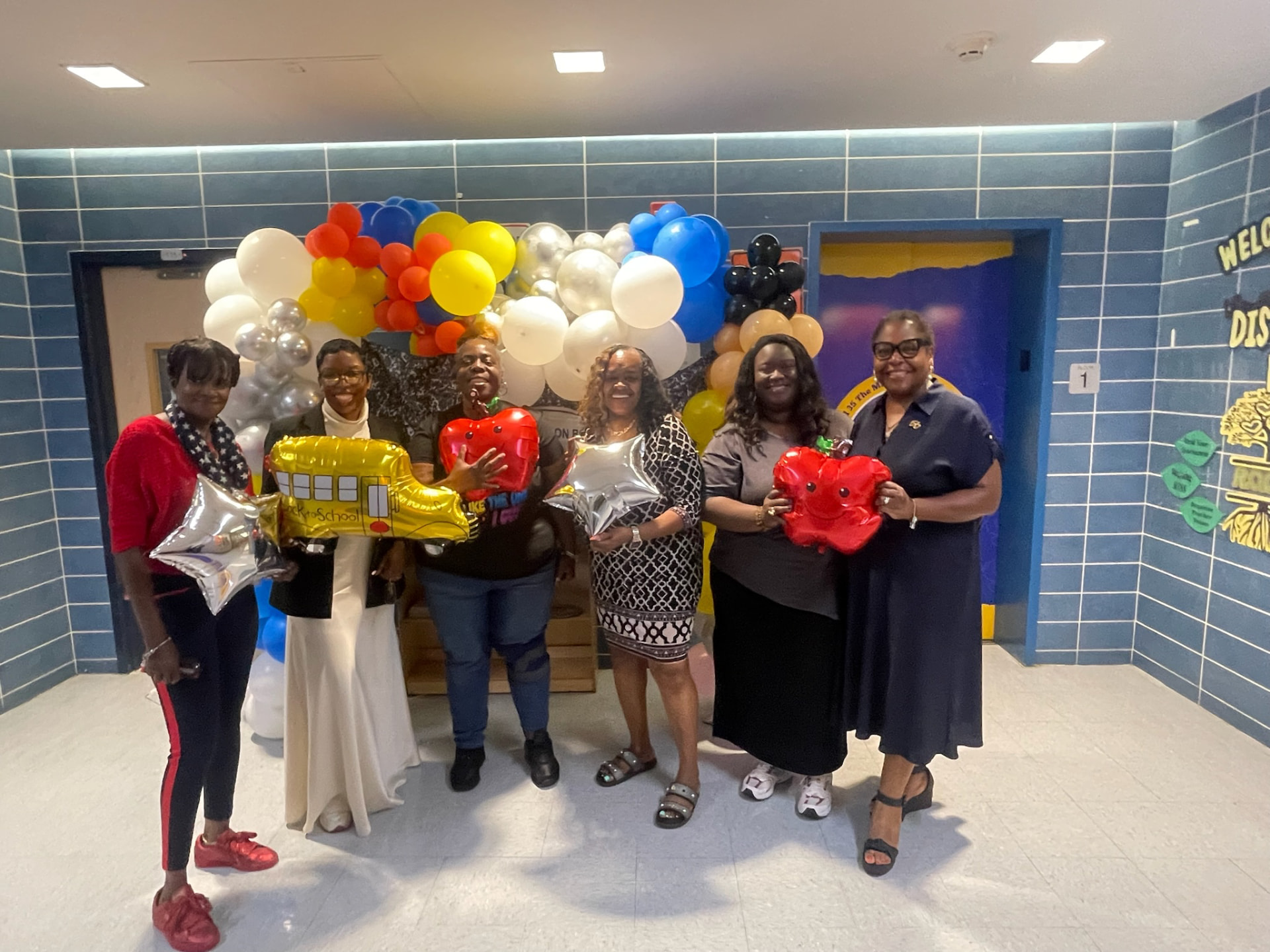 Six people holding colorful balloons in a tiled hallway